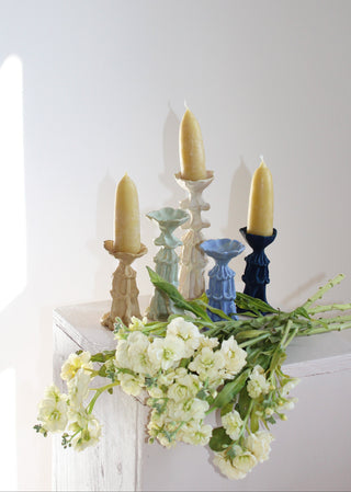 Four decorative taper candles in different muted colors displayed on a white pedestal with white flowers in the foreground against a white wall.